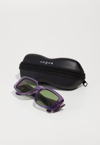 Πετρόλ ορθογώνια γυαλιά ηλίου με πράσινους φακούς, με παχύ σκελετό, βρίσκονται δίπλα σε μία μαύρη σκληρή θήκη με φερμουάρ και άσπρο λογότυπο.