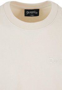 T-shirt en coton beige avec un col rond et un petit logo brodé. Le tissu a une texture lisse et les coutures sont finies avec soin.