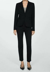 Costume noir sur mesure comprenant un blazer cintré avec un bouton unique et deux poches avant, associé à un pantalon slim et des talons noirs pointus.