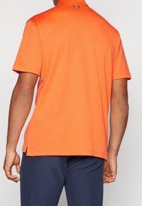 Oranžové polo tričko s krátkým rukávem a texturovaným vzorem, se zakřiveným spodním lemem a malým logem značky na zadním výstřihu. V kombinaci s tmavými kalhotami.