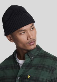 Un berretto in maglia a coste nera sopra la testa di un uomo, abbinato a una camicia a scacchi verde e nera in flanella con un piccolo logo giallo sul petto.