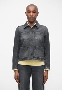 Grijze spijkerjas met twee borstzakken, klassiek kraag, knoopsluiting en een geribbelde gele trui eronder. Beschikt over een recht lijfje.