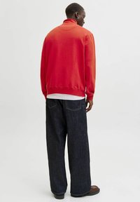 Felpa rossa con collo alto, vestibilità ampia, abbinata a jeans larghi scuri, con cuciture a contrasto e una texture morbida. Scarpe nere visibili.