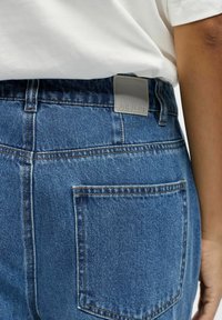 Gros plan d'une personne portant un jean en denim bleu avec une poche arrière carrée et une étiquette grise sur la ceinture, associée à une chemise blanche.