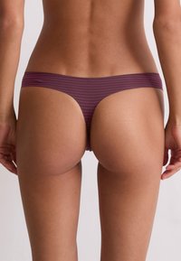 Rear view of a person wearing purple and black striped thong underwear against a plain light background.