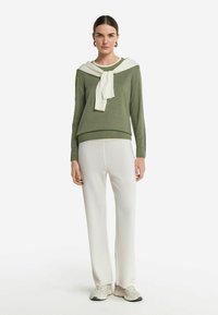 Oliwkowy sweter z białym szalikiem związanym przy dekolcie, w zestawieniu z białymi spodniami o szerokich nogawkach i beżowymi butami sportowymi. Gładki materiał i luźny krój.