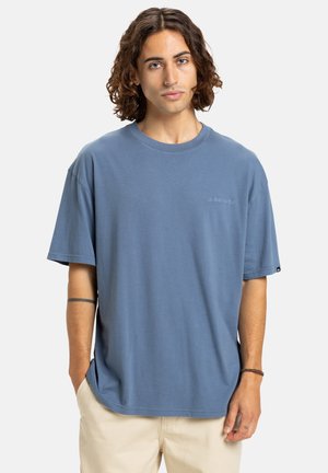 Young person with curly brown hair wearing a blue oversized t-shirt and beige pants, standing with one hand in pocket against a white background.