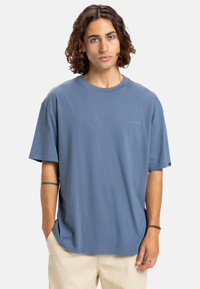 Ung person med krøllet brunt hår iført en blå oversized T-shirt og beige bukser, der står med den ene hånd i lommen mod en hvid baggrund.