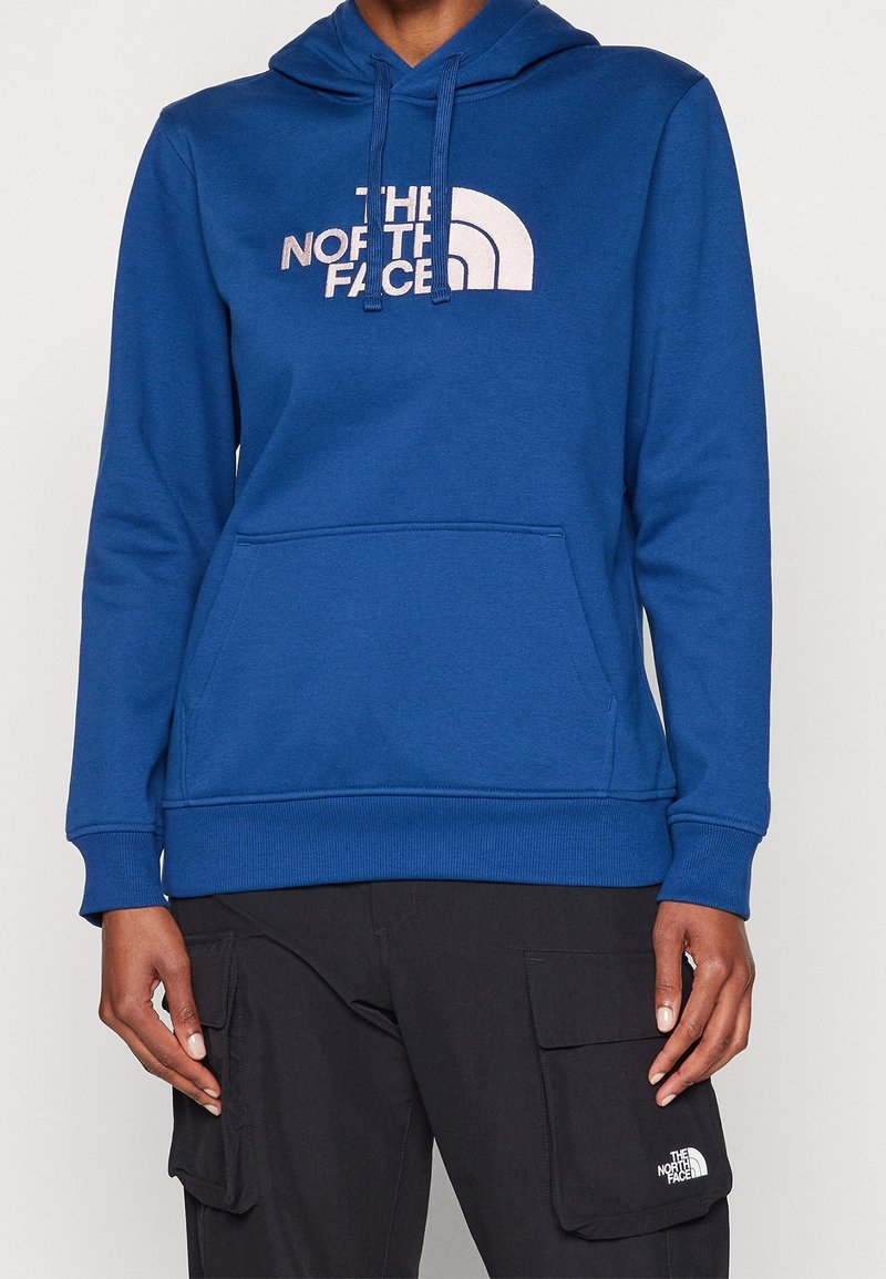 Sweat à capuche bleu avec logo blanc "The North Face", poche avant, porté avec un pantalon cargo noir affichant un petit logo blanc sur la poche.