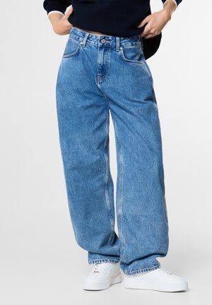 Persoon draagt lichtblauwe wijde jeans met witte sneakers en een donker lange mouwen top, handen rusten op de heupen.