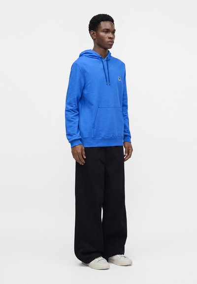 Jeune homme debout portant un sweat à capuche bleu vif, un pantalon large noir et des baskets blanches sur un fond clair uni.