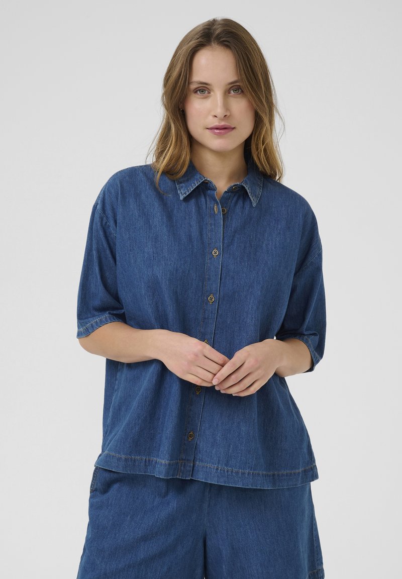 Denim shirt with a collared neckline, short sleeves, and a relaxed fit. Features gold-tone buttons and contrasting stitching details.