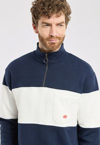 Pull en bleu marine et blanc avec un col zippé à mi-hauteur, comportant une poche avant et un tissu texturé, mettant en valeur un design coloré.