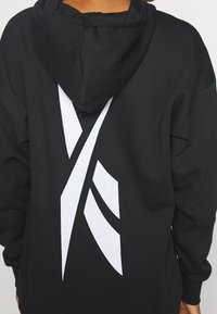 Czarna bluza z kapturem wykonana z bawełny, z dużym białym geometrycznym wzorem na plecach. Kapturek jest regulowany, a mankiety są dziane.