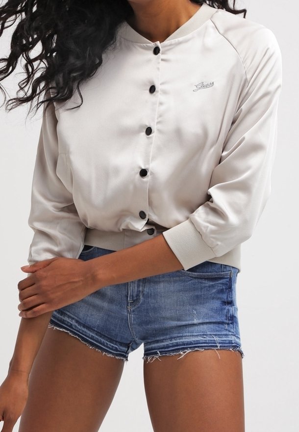 Veste bomber en satin beige clair avec un col rond, des boutons noirs, des poignets côtelés et un logo sur le devant. Associée à un short en denim déchiré.