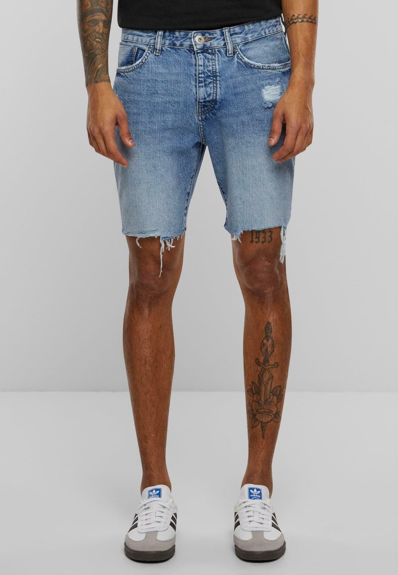 Svetlomodré denim šortky s rozstrapkaným lemom, piatimi vreckami a jemným opotrebovaním. Skombinované so šedými a bielymi športovými topánkami.