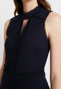Tmavě modré bezramínkové šaty s vysokým límcem, s černou krajkovou ozdobou podél výstřihu a pasu, a jemným průstřihem vpředu.