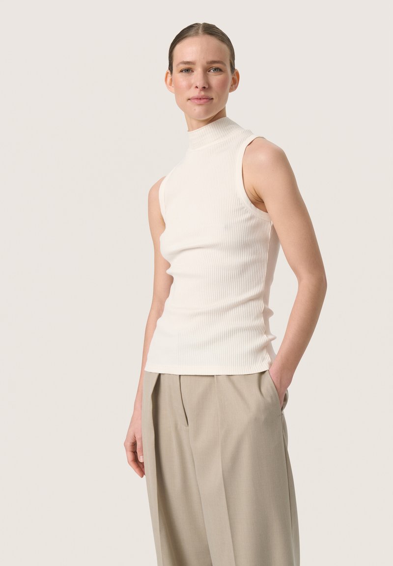Weißes, geripptes, ärmelloses Rollkragenoberteil mit tailliertem Design, kombiniert mit weit geschnittenen beigen Hosen. Glatte Textur, klare Linien, minimalistischer Stil.