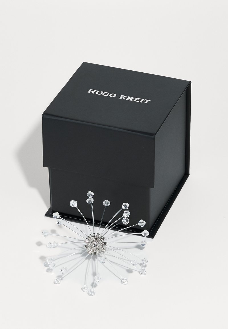 Sort firkantet boks med "Hugo Kreit" i hvid. Sølv metallisk solstrålingsdesign med klare krystal detaljer, der stråler udad.