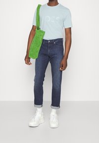 Světle modré tričko s potiskem "LOSS", tmavě modré džíny, zelená látková taška přes rameno a bílé tenisky s texturovaným povrchem.