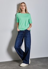 Žena s dlouhými blond vlasy, oblečená v zeleném tričku s krátkým rukávem, širokých tmavě modrých džínách a bílých teniskách, stojí proti šedé zdi.