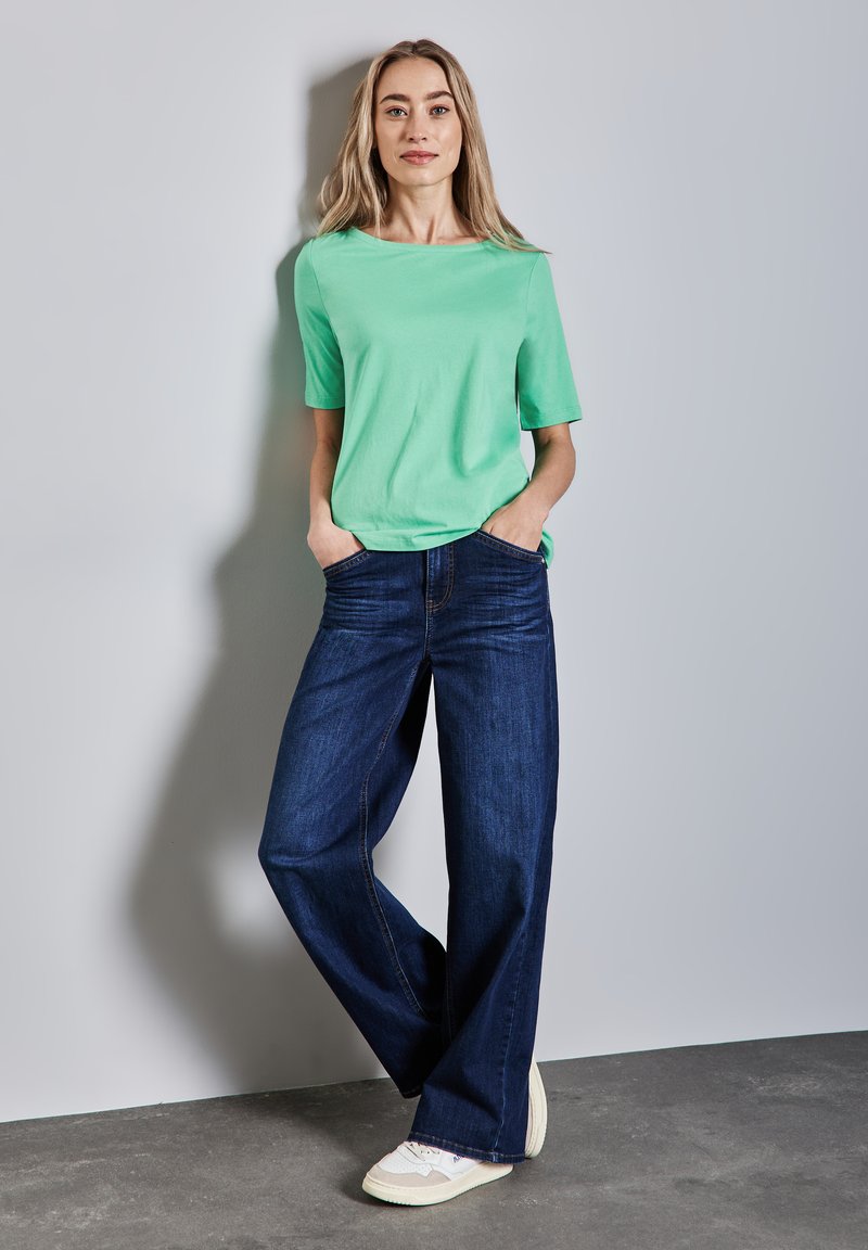 Žena s dlouhými blond vlasy, oblečená v zeleném tričku s krátkým rukávem, širokých tmavě modrých džínách a bílých teniskách, stojí proti šedé zdi.