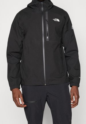 Osoba nosząca czarną kurtkę z kapturem The North Face z kieszeniami na zamek i logo na piersi i rękawie.
