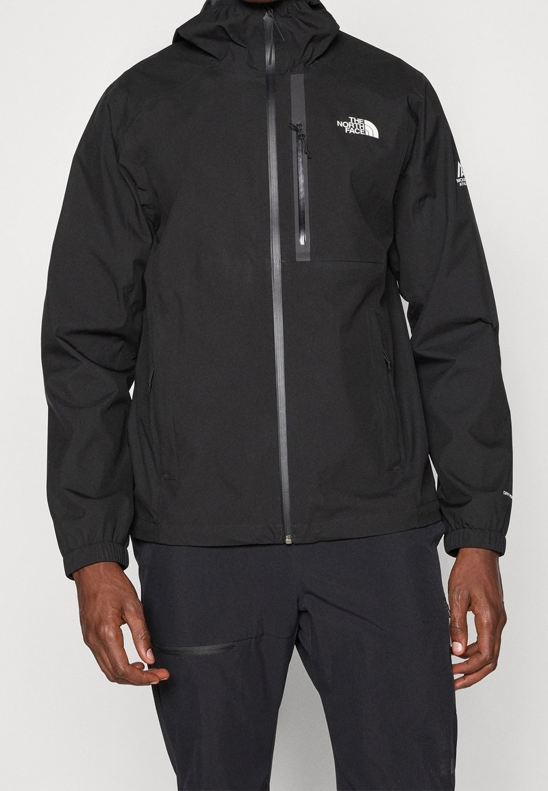 Personne portant une veste noire à capuche The North Face avec des poches zippées et un logo sur la poitrine et la manche.