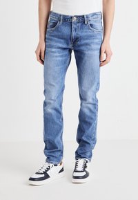 Person klädd i ljusblå slim-fit jeans och svart-vita sneakers, stående mot en enfärgad vit bakgrund.