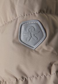 Bē�žs pūkains jaka audums ar gludu tekstūru, uz kura virsmas ir izšūts pelēks sešstūru logotipa uzlīme.