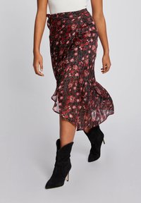 Jupe portefeuille noire à motifs floraux rouges et blancs, en tissu transparent, avec un ourlet volants et un nœud sur le côté, associée à des bottines en daim noires.