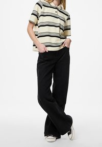 En person står med en hand i fickan, iförd en randig kortärmad skjorta och svarta vida byxor, mot en enkel vit bakgrund.