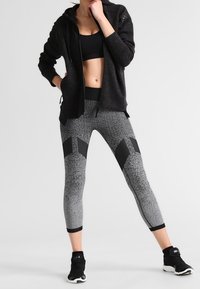 Sweat à capuche noir zippé avec finition texturée, associé à des leggings gris ajustés présentant un motif tacheté et des accents géométriques noirs. Baskets noires.