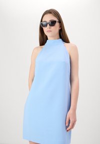 Svetlo modré šaty s halter krkom vyrobené z hladkej tkaniny, majú priliehavý korzet a rozšírenú sukňu. Modelka má na sebe tmavé slnečné okuliare.