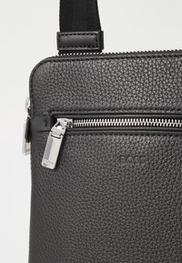 Sac en cuir noir texturé avec une fermeture éclair argentée, une étiquette de marque et une bandoulière réglable. Design compact et rectangulaire adapté à un usage quotidien.