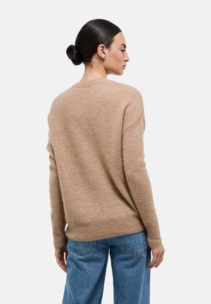 Femme aux cheveux noirs coiffés en chignon, portant un pull en maille beige et un jean bleu, de profil avec une expression neutre sur un fond blanc.