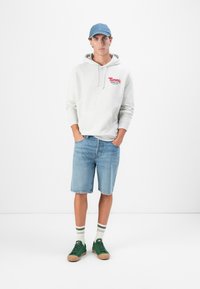 Lys grå hættetrøje med rød tekst, blå denim shorts, grønne sneakers med brune såler og stribede sokker. En simpel, afslappet påklædning.