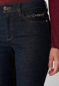 Ciemnoniebieskie dżinsy z denimu z pomarańczowym przeszyciem, akcentem łańcuszka przy przedniej kieszeni oraz klasycznym, pięciokieszeniowym krojem. Gładka textura.