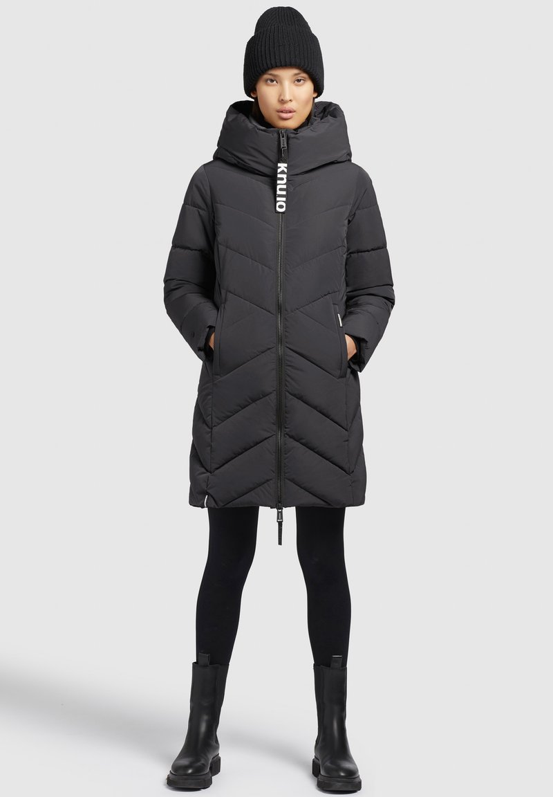 Doudoune noire avec couture en chevrons, col haut et zip frontal. Portée sur des leggings noirs et des bottes noires épaisses, complétée par un bonnet en tricot.