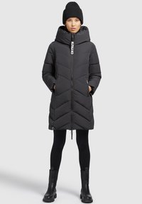Zwarte gewatteerde jas met chevronpatroon, hoge kraag, ritsluiting en zijzakken. Gecombineerd met zwarte leggings en enkellaarsjes.