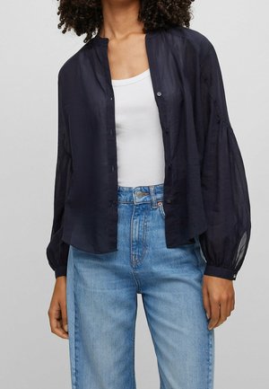 Kvinde iført mørkeblå knap-op-bluse med puffede ærmer over hvid tanktop og lyseblå højtaljede jeans.