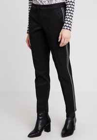 Černé kalhoty s úzkým střihem, s bílými bočními pruhy, hladkou texturou a středně vysokým pasem. Nosené s černými kotníkovými botami na podpatku.