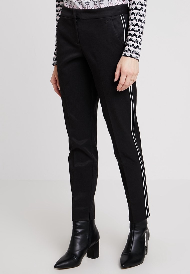 Černé kalhoty s úzkým střihem, s bílými bočními pruhy, hladkou texturou a středně vysokým pasem. Nosené s černými kotníkovými botami na podpatku.