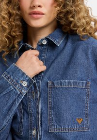Jeanshemd in verblasstem Blau mit einer Brusttasche, silbernen Knöpfen, kontrastierenden Nähten und einem kleinen Herz-Logo-Detail auf der Tasche.