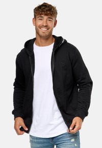 Sort zip-tætsiddende hoodie med en frontlomme, justerbar hætte og et lille logo nær brystet. Iført over en hvid t-shirt med blå jeans.