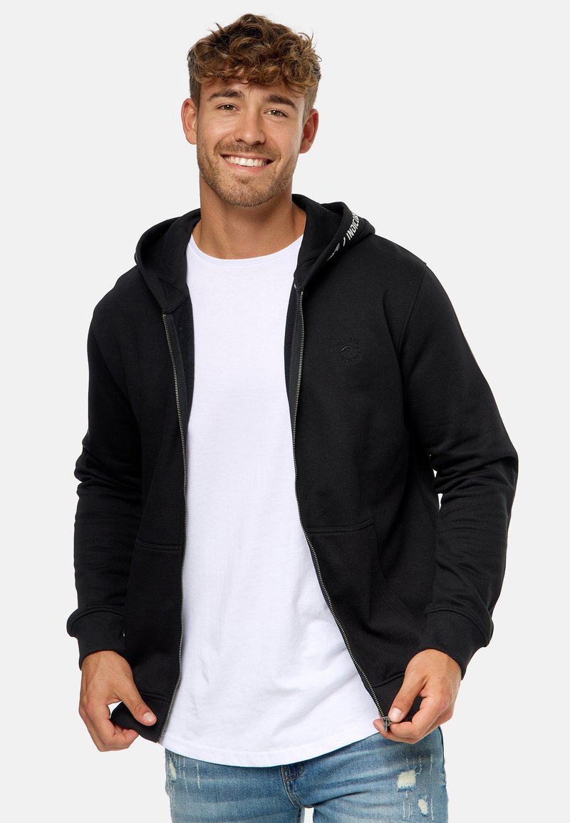 Sort zip-tætsiddende hoodie med en frontlomme, justerbar hætte og et lille logo nær brystet. Iført over en hvid t-shirt med blå jeans.