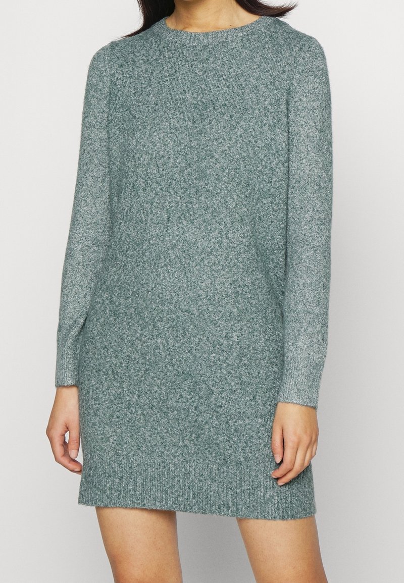 Femme portant une robe-pull en maille grise texturée, manches longues, longueur genou, debout devant un fond clair uni.