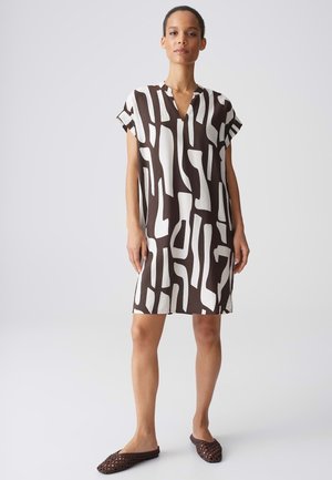 Donna in piedi con un vestito a maniche corte dal motivo astratto bianco e nero e scarpe slip-on intrecciate marroni su uno sfondo semplice.