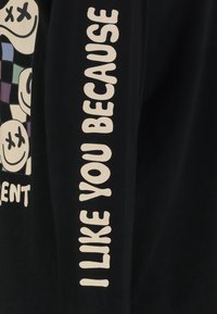 Schwarzes Langarmshirt mit fettem weißen Text "ICH MAG DICH, WEIL" auf dem Ärmel und bunten grafischen Designs auf der Vorderseite.
