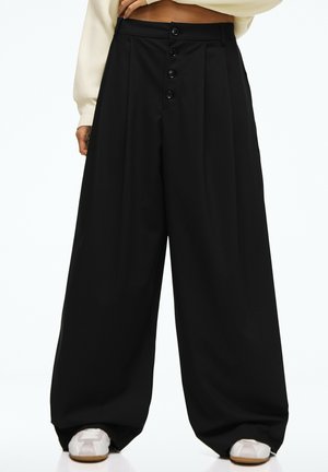 Pantalon noir à jambes larges avec quatre boutons sur le devant, porté avec des baskets blanches et un pull court crème.